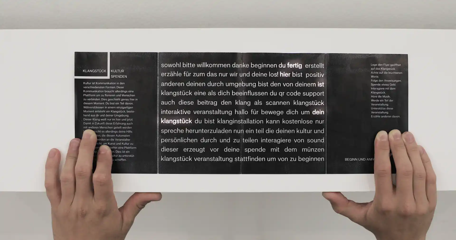 A photo of the installation Klangstück from above. A persons hands are visible, the person is holding a paper flyer on the slanted face of the installation. The flyer is black with white font printed on it. The middle displays a seemingly random amount of words. Some of them are illuminated through the back of it creating a sentence.