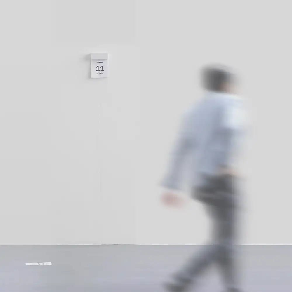 a wall calendar hanging on a white wall with one calendar page laying on the floor. there is a person walking in front of it